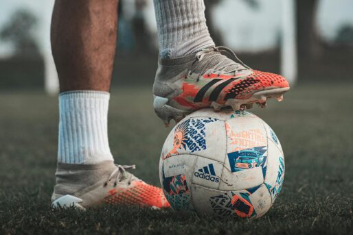 Close-up of a soccer player wearing Adidas cleats, holding a branded soccer ball on grass.